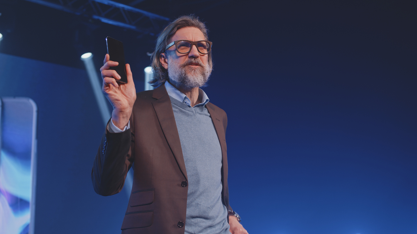 A mature creator in a casual suit on stage, holding a modern mobile phone and showing the release and speaking about the new device in a large room 
